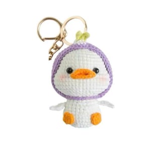 Puede incluir: Un llavero de pato hecho a mano con ganchillo. El pato es blanco con pico y patas naranjas, y un sombrero morado. El llavero tiene un anillo de metal dorado.