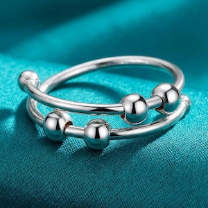 May include: A silver-colored fidget ring with a double band design. The ring features three small, shiny, spherical beads that can be moved along the band. The ring is displayed on a teal-colored, textured surface.