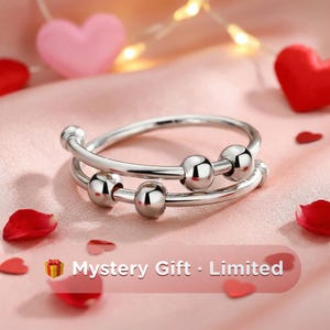 May include: A silver-colored ring with three small, shiny spheres. The ring has a double-band design. The background includes pink fabric, red heart-shaped decorations, and the text "Mystery Gift - Limited".