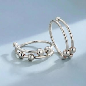 May include: Two silver-colored fidget rings with a double-band design. Each ring features small, spherical beads that can be moved along the band. The rings are displayed against a light blue background.
