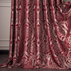 Può includere: Tenda bordeaux con un motivo dorato ripetuto. Il design presenta forme floreali e geometriche stilizzate. Il tessuto ha una leggera lucentezza, che ne esalta l'aspetto lussuoso. La tenda è drappeggiata, mostrando la sua consistenza.