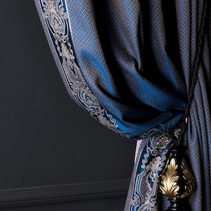 Op de afbeelding: Blauw gordijn met patroon en een decoratieve rand met een sierlijk ontwerp in zilver en wit. Een zwarte en gouden kwast is aan het gordijn bevestigd, wat een vleugje elegantie toevoegt. Het gordijn is gedrapeerd tegen een donkergrijze muur.