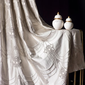 Puede incluir: Una lujosa cortina blanquecina con intrincados bordados plateados y un diseño fluido. Dos jarrones de cerámica blanca con detalles dorados se asientan sobre una mesa cubierta con la cortina. El patrón de la cortina incluye formas ornamentadas y detalles en forma de cadena.