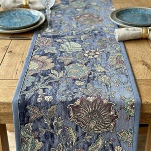 May include: A blue floral table runner with a light blue border. The runner displays a detailed pattern of flowers in shades of blue, green, and burgundy. The runner is on a wooden table, with place settings visible.