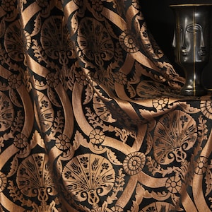 May include: Close-up of a draped fabric with a repeating pattern of stylized floral and geometric shapes in black and bronze. The fabric has a luxurious, textured appearance, with folds and shadows adding depth. A dark vase with a face design is on the right.