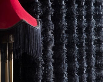 Puede incluir: Primer plano de una cortina negra texturizada con un diseño vertical con flecos. Una pantalla de lámpara roja con flecos negros y una base de lámpara dorada son visibles. La cortina tiene una apariencia suave y afelpada.