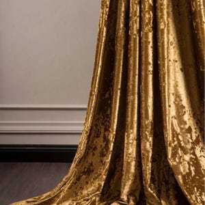 May include: A gold velvet curtain with a textured finish. The fabric is draped, creating folds and shadows. The curtain is set against a neutral wall and dark floor, highlighting its rich colour and texture. The overall effect is one of opulence.