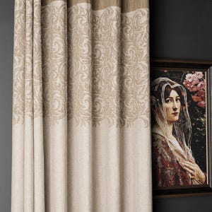 Può includere: Pannelli di tenda beige e marrone chiaro a fantasia con occhielli. Le tende presentano un motivo floreale dettagliato nella parte inferiore e una fascia marrone chiaro in tinta unita nella parte superiore. Un dipinto incorniciato è appeso alla parete a destra.