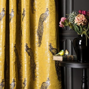 May include: A mustard yellow curtain featuring a repeating pattern of black and gold peacocks. The fabric has a subtle sheen and a textured appearance. The peacock design is detailed with visible feathers and crests, arranged in vertical columns across the fabric.