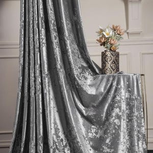 Op de afbeelding: Zilveren crushed velvet gordijn met een verweerd patroon. Het gordijn is gedrapeerd en toont zijn textuur en glans. Een decoratieve vaas met bloemen staat op een tafel bedekt met dezelfde stof, wat een samenhangend geheel creëert.