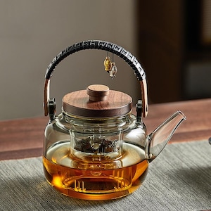 May include: A clear glass teapot with a wooden lid and handle, filled with amber-colored tea. The handle is wrapped in black material and has small gold-colored fish charms hanging from it. The teapot has a built-in infuser.