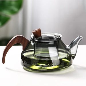 Puede incluir: Una tetera de vidrio transparente con asa y tapa de madera. La tetera tiene un infusor incorporado y está llena de un líquido verde. El asa y la tapa son de madera marrón oscuro. La tetera está sobre una superficie blanca.