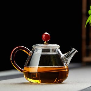 Puede incluir: Una tetera de vidrio transparente con asa y boquilla marrones. La tetera tiene un pomo rojo en la tapa y está llena de té. La tetera está sobre una superficie gris.