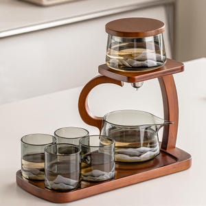 May include: A wooden tea set with a dark brown finish. The set includes a glass teapot, four glasses, and a tea infuser. The glasses and teapot have a mountain design. The set is on a wooden tray.