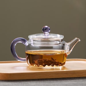 Op de afbeelding: Een heldere glazen theepot met een paarse handgreep en deksel. De theepot is gevuld met gezette thee en staat op een houten dienblad. De theepot heeft een afgeronde body en een tuit.