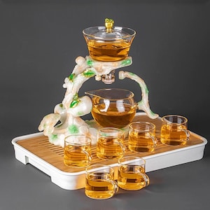 Può includere: Un set da tè con una teiera in vetro trasparente, sei piccole tazze da tè e un supporto decorativo. Il supporto è bianco e verde con un design a forma di ramo. Il tè è color ambra e il set poggia su un vassoio di bambù.