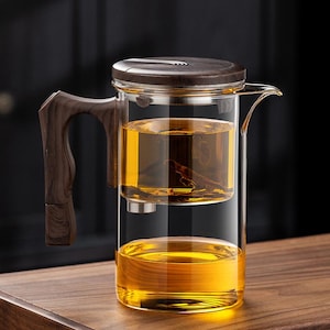 Può includere: Una teiera in vetro trasparente con manico, coperchio e infusore in legno. La teiera è riempita di tè color ambra. Il manico e il coperchio sono di colore marrone scuro. La teiera è su una superficie di legno.