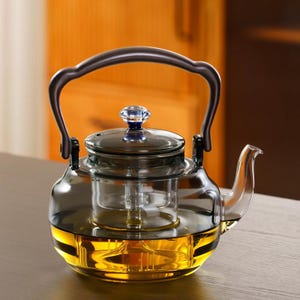 May include: A clear glass teapot with a dark handle and lid. The teapot has a built-in infuser and is filled with a golden liquid. A blue gem tops the lid. The teapot is sitting on a wooden surface.