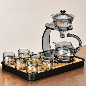 Può includere: Un set da tè in vetro con teiera, infusore e sei tazze. La teiera e le tazze hanno un design decorativo. Il set è su un vassoio nero con un inserto in bambù e un supporto trasparente.