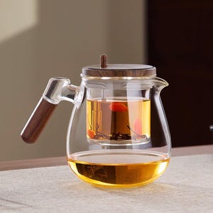 Puede incluir: Una tetera de vidrio transparente con asa y tapa de madera. La tetera está llena de té y tiene un infusor incorporado. El té es de color dorado y la tetera está sobre una superficie clara.