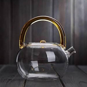 Puede incluir: Una tetera de vidrio transparente con cuerpo redondeado y asa dorada. El pico y la tapa también son de vidrio transparente. El asa es arqueada y se conecta a la parte superior de la tetera. La tetera está sobre una superficie de madera oscura.