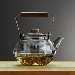 Może przedstawiać: Przezroczysty szklany czajnik z teksturowanym korpusem i drewnianym uchwytem. Czajnik posiada dzióbek i pokrywkę z drewnianym guzikiem. Uchwyt jest przymocowany złotym metalem. Czajnik jest wypełniony złotym płynem.