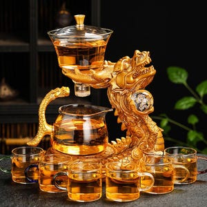 Könnte beinhalten: Ein goldenes Drachen-Teeservice mit einer Glaskanne, einem Glas-Serviergefäß und mehreren kleinen Glasteetassen. Das Teeservice ist mit bernsteinfarbenem Tee gefüllt. Der Drache ist aufwendig detailliert.