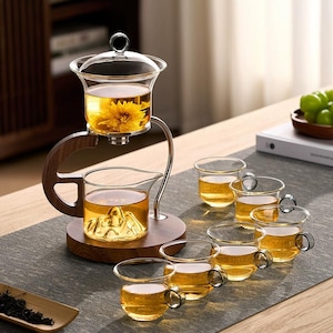 Puede incluir: Un juego de té de vidrio con base y asa de madera. El juego incluye una tetera con una flor en su interior, una jarra con un diseño de montaña y seis tazas de té pequeñas. El té es de color ámbar.