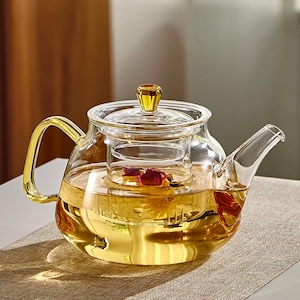 Può includere: Una teiera in vetro trasparente con manico, coperchio e infusore dorati. La teiera è riempita con un liquido color oro e contiene petali di rosa essiccati. La teiera è appoggiata su una superficie marrone chiaro.