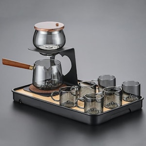 Può includere: Un moderno set da tè con una teiera in vetro con manico in legno, un infusore in vetro con coperchio in legno e sei tazze da tè in vetro abbinate. Il set è presentato su un vassoio nero con un inserto in bambù.