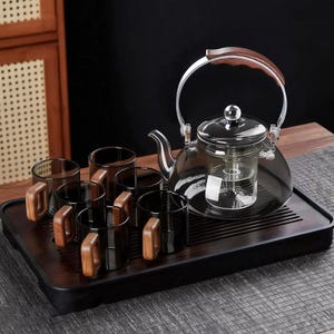 May include: A clear glass teapot with a wooden handle and a set of six matching cups with wooden handles. The tea set is displayed on a dark wooden tray, perfect for serving tea. The teapot has a built-in infuser.