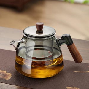 Può includere: Una teiera in vetro trasparente con coperchio in acciaio inossidabile e manico in legno. La teiera è riempita di liquido color ambra, probabilmente tè. Il manico e il pomello del coperchio sono in legno.