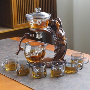 Puede incluir: Juego de té con una tetera de vidrio, un cuenco de servir de vidrio y seis tazas de té pequeñas de vidrio. La tetera y el cuenco de servir tienen diseños florales. El juego está sostenido por una escultura de madera de una mujer. El té es de color ámbar.