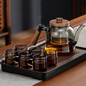 May include: A glass teapot with a wooden lid and handle, and six small glass teacups, presented on a dark wooden tray. The teapot has an infuser and is filled with amber liquid. The set is designed for tea.