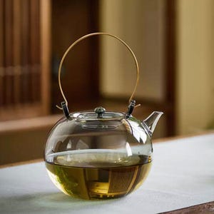 Può includere: Una teiera in vetro trasparente con corpo rotondo e manico dorato. La teiera è riempita con un liquido chiaro, probabilmente tè. Il coperchio è argentato con un pomello decorativo. La teiera è su una superficie bianca.