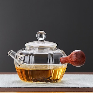 May include: A clear glass teapot with a wooden handle and lid. The teapot is filled with tea, and the handle is a dark brown color. The teapot is sitting on a light-colored surface.