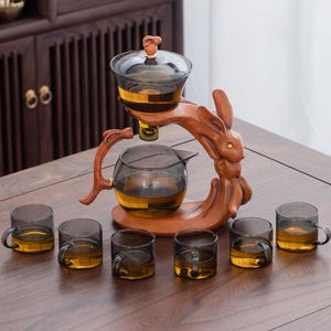 Könnte beinhalten: Ein Teeservice mit einem hölzernen Hasen-Ständer, der eine Glaskanne und einen Tee-Infuser hält. Sechs kleine Teetassen mit Griffen sind davor angeordnet. Der Tee hat eine bernsteinfarbene Farbe. Das Set steht auf einem Holztisch.