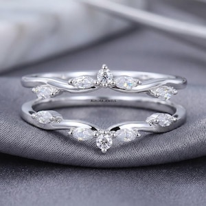 May include: A silver-toned ring guard featuring two curved bands that meet at a central point. Each band is adorned with marquise-cut and round-cut clear stones arranged in a symmetrical pattern. The text AURORA JEWELS is printed in the center.