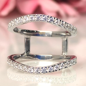 May include: A silver ring with two curved bands, each lined with small, clear gemstones. The ring is connected by two vertical bars. The text "925" is visible on the ring. The words "AURORA JEWELS 6" are also visible.