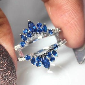 May include: A silver ring guard with a crown design, featuring marquise and round-cut blue gemstones. The ring is held between fingers, showcasing the intricate details of the jewelry.