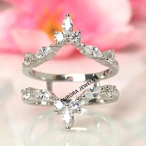 Puede incluir: Un protector de anillo plateado con un diseño en forma de V, adornado con múltiples piedras preciosas claras y brillantes. El protector de anillo está diseñado para colocarse junto a otro anillo. El texto "AURORA JEWELS 6" es visible.