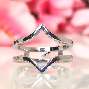 Puede incluir: Un anillo plateado con un diseño único en forma de V. El anillo presenta dos bandas, una encima de la otra, cada una con una parte superior puntiaguda. El texto "AURORA JEWELS 6" es visible. El fondo es rosa.