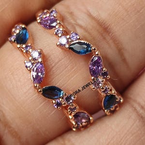 May include: Two rose gold rings, each adorned with a series of blue, purple, and clear gemstones. The gemstones are arranged in a floral or leaf-like pattern, creating a delicate and elegant design. The rings are set against a neutral background.