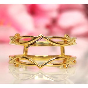 May include: A gold-colored ring guard with an openwork design. The ring guard features a decorative pattern with a V-shaped element and the text "AURORA JEWELS 6". The ring guard is designed to be worn with another ring.