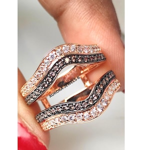 Può includere: Due anelli in oro rosa con un design ondulato. Ogni anello presenta una fila di piccole pietre chiare e una fila di pietre marrone scuro. Gli anelli sono tenuti dalle dita di una persona. Il testo "AURORA JEWELS 6" è visibile.