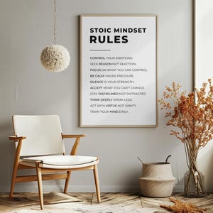 Może przedstawiać: Oprawiony wydruk z napisem "STOIC MINDSET RULES" wisi na szarej ścianie. Obok znajduje się drewniane krzesło z białym siedziskiem. Widoczna jest r&oacute;wnież dekoracyjna lampa wisząca i wazon z suszonymi roślinami.