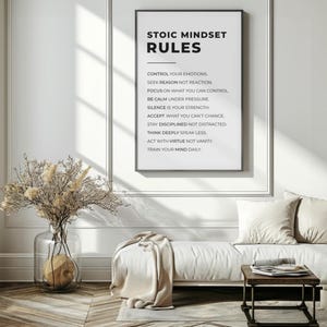 Może przedstawiać: Oprawiony wydruk z czarnym tekstem "STOIC MINDSET RULES". Zasady są wymienione poniżej tytułu. Biała sofa z poduszkami i mały drewniany stolik znajdują się przed wydrukiem. Po lewej stronie wazon z suszonymi kwiatami.