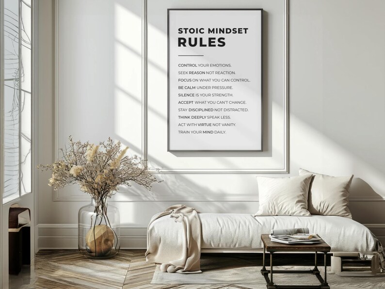 Może przedstawiać: Oprawiony wydruk z czarnym tekstem "STOIC MINDSET RULES". Zasady są wymienione poniżej tytułu. Biała sofa z poduszkami i mały drewniany stolik znajdują się przed wydrukiem. Po lewej stronie wazon z suszonymi kwiatami.