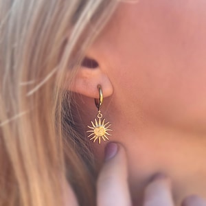 May include: Gold-colored hoop earring with a sun pendant. The sun pendant features a smiling face and radiating lines. The earring is worn on the ear. The image is well-lit and the earring is in focus.
