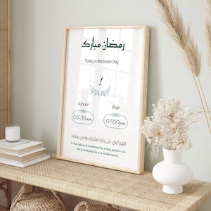 Puede incluir: Una impresión enmarcada con un marco de madera clara muestra texto en árabe e inglés, incluyendo "Ramadan Mubarak" y "Today is Ramadan Day". La impresión presenta un horario para Suhoor e Iftar, con los horarios indicados. Un jarrón blanco con flores está cerca.